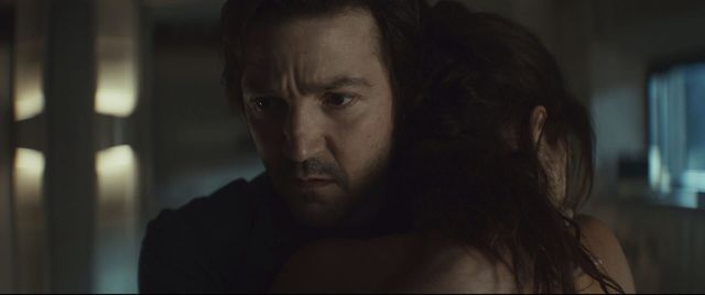 A man with stubble embraces a woman with long dark hair in a dimly lit room, his expression tense and distant.