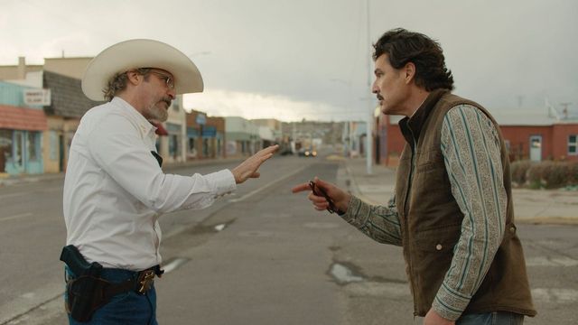 Two men face each other on a small-town street; one wears a cowboy hat, glasses, and holstered gun, the other a vest over a striped shirt.