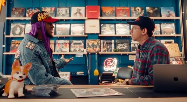 Two men talking at a record store counter, one in a denim jacket with purple durag, the other in plaid shirt and cap.