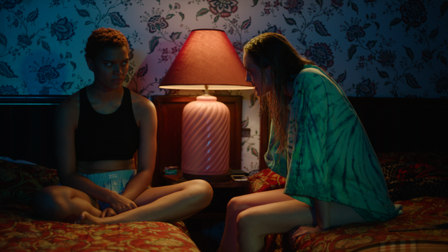 Two young women sit on a bed in a dimly lit room with floral wallpaper, a pink lamp glowing between them on a nightstand.