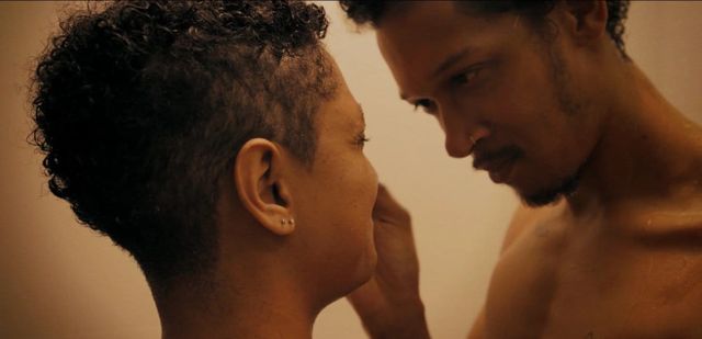 Two shirtless people facing each other in close intimacy, bathed in warm golden light, with curly hair and small earring.