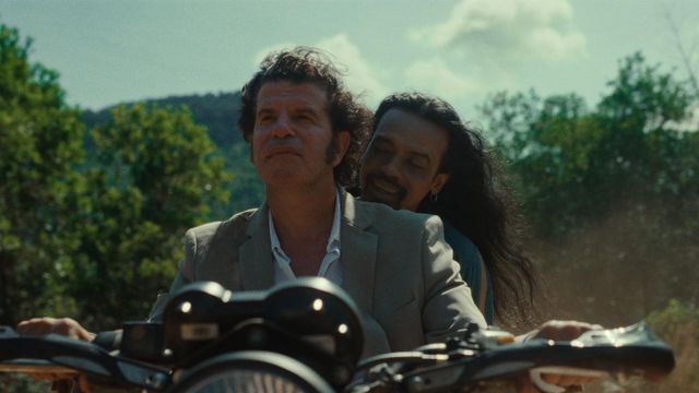 Two men riding a motorcycle outdoors, the driver in a gray blazer, the passenger with long dark hair smiling behind him.