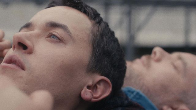 Close-up of two young men lying back, heads together, one gazing upward with blue eyes, the other eyes closed, outdoors.