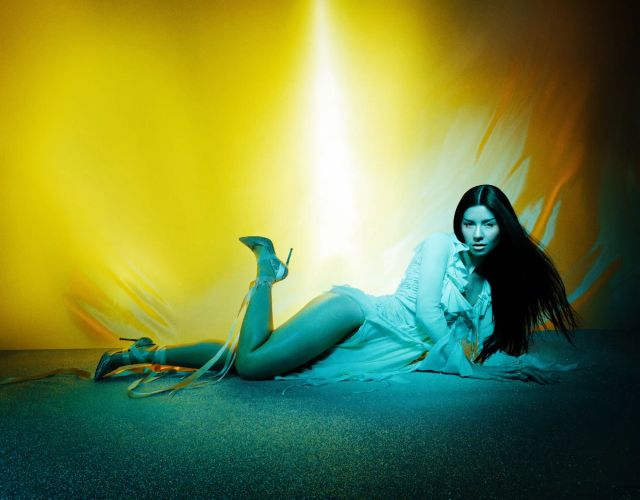 Woman with long dark hair reclining on floor in white ruffled dress and strappy high heels, lit by teal and yellow lights.