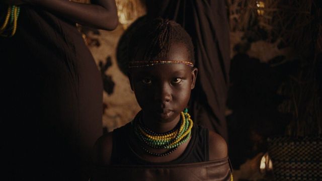 Young African child with braided hair, beaded headband, and layered green and yellow bead necklaces in a dimly lit space.