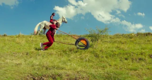 Person in red tracksuit running downhill pulling a flaming tire on ropes, while another person rides a white horse behind them on green grass.