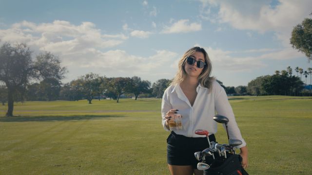 Woman in white blouse, black shorts, and sunglasses holding a drink beside a golf bag on a sunny golf course.