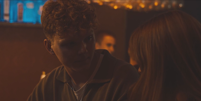 Young person with curly reddish-blond hair in a zip-up hoodie facing someone with straight blonde hair in dim warm lighting.