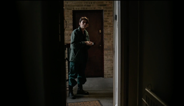 Woman in dark green jumpsuit and boots standing in a brick-walled hallway near a brown door, viewed through a dark doorframe.
