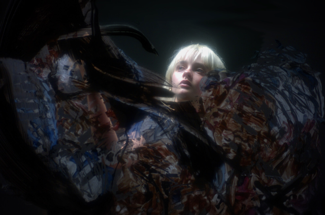 Young woman with platinum blonde bob haircut surrounded by abstract swirls of dark paint-like textures in moody lighting.