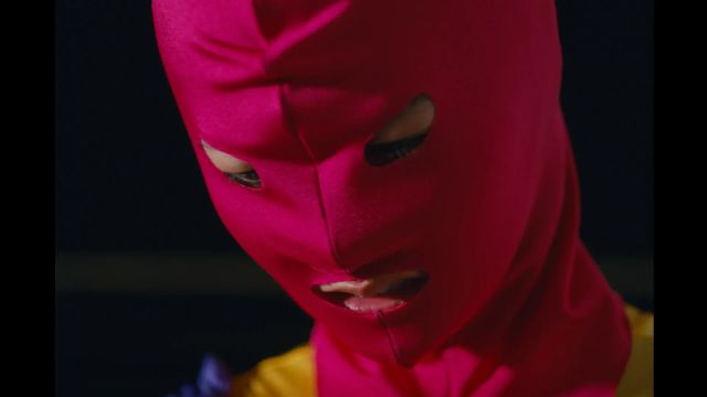 Close-up of a person wearing a bright pink ski mask/balaclava with eye and mouth openings, dressed in yellow clothing.