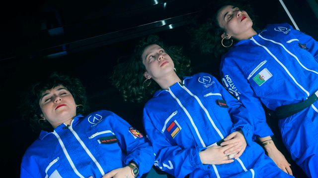 Three women in blue astronaut-style jumpsuits with patches and flags lie side by side with eyes closed against a dark background.
