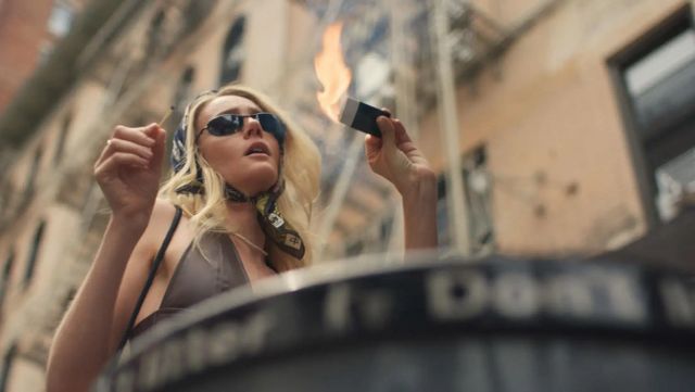 Blonde woman in sunglasses and scarf holds a burning piece of paper over a metal trash can on an urban street.