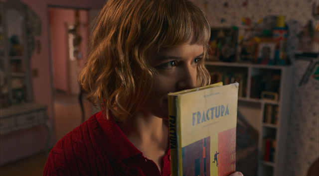Person with shoulder-length reddish-brown hair in a red sweater holding a book titled "Fractura" near their face in a colorful room.