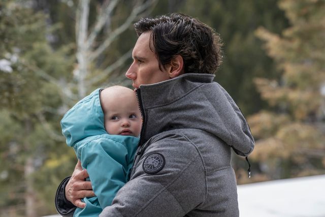 Man in gray Canada Goose jacket holding a baby in a light blue snowsuit outdoors with trees in the background.