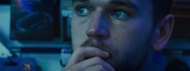 Close-up of a young man with stubble resting his hand on his chin, looking upward thoughtfully, lit by blue light.