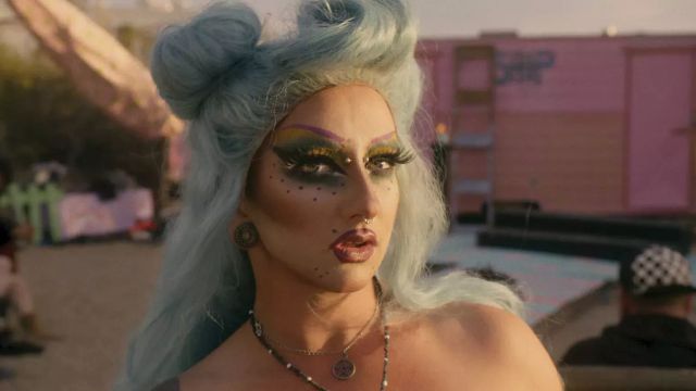 Drag performer with light blue upswept hair, bold yellow-green eyeshadow, purple brows, dotted cheek art, and septum ring outdoors.