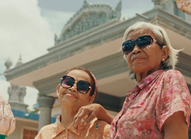 Two elderly women wearing sunglasses and floral shirts smile in front of an ornate white temple on a sunny day.