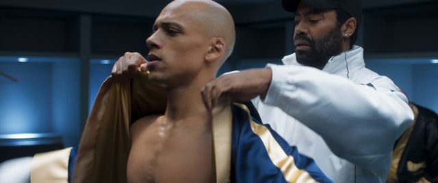 Bald shirtless man being helped into a blue and gold boxing robe by a bearded man in a white jacket and cap in a locker room.