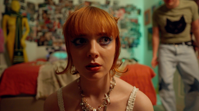 Young woman with orange bangs and dramatic blue eye makeup wearing a cross necklace, in a colorful bedroom with people behind her.