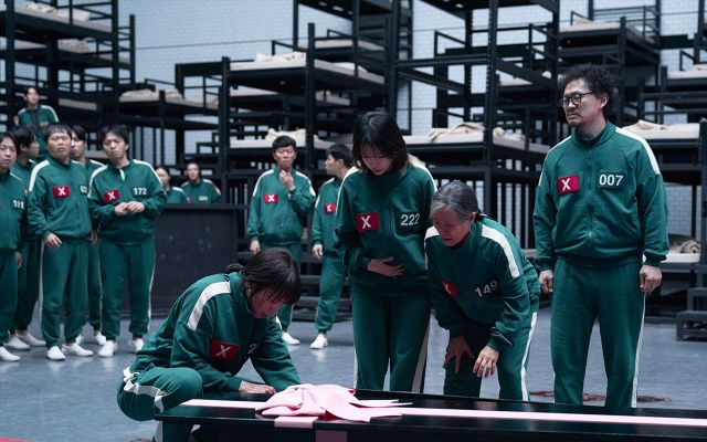 Squid Game scene: players in green numbered tracksuits gather around a coffin with pink cloth in the dormitory set.