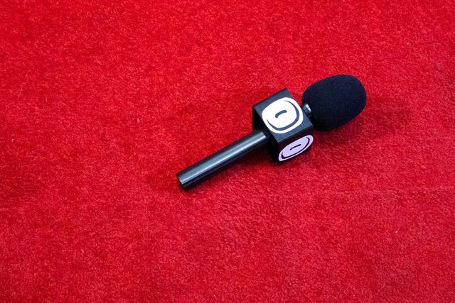 Black broadcast microphone with foam windscreen and logo cube on red carpet surface.