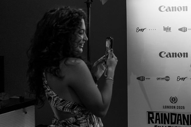 Woman with curly hair taking a phone photo at the Raindance Film Festival London 2025, standing near a sponsor backdrop.