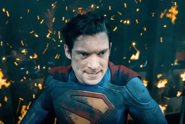 Man in a dirty, battle-damaged Superman suit grimacing with sparks and flames flying in the background behind him.