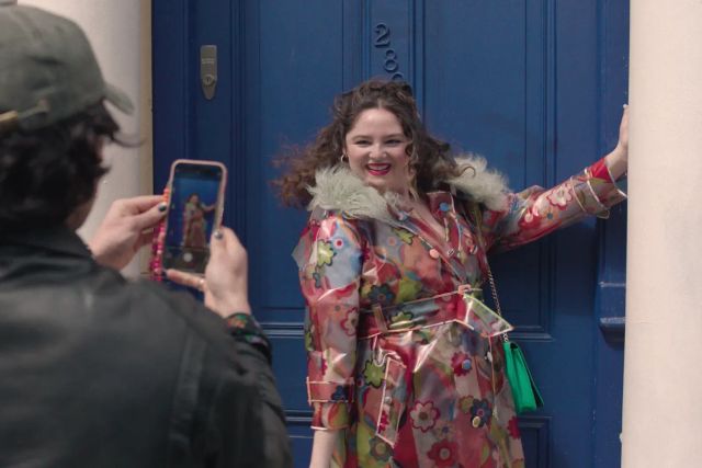 Smiling woman in colorful floral trench coat with feather collar poses by a blue door while someone photographs her with a phone.
