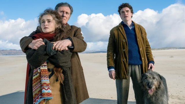 Three people on a windswept beach: a woman in a red scarf embraced by an older man in a brown coat, a young curly-haired man with a large shaggy dog.