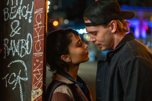 Two young people lean close as if about to kiss on a nighttime city street beside a graffiti-covered pole.
