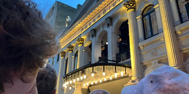 Illuminated facade of the London Palladium theatre at dusk, viewed from below with curly-haired person in foreground.