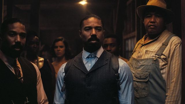 Three men stand in a dimly lit room; the center man wears a vest, blue shirt, and holster, flanked by others in period clothing.