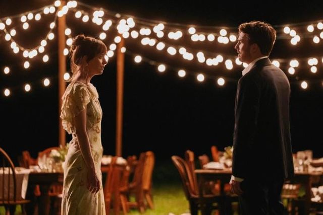 Two people facing each other at night under string lights at an outdoor event with dining tables and chairs behind them.