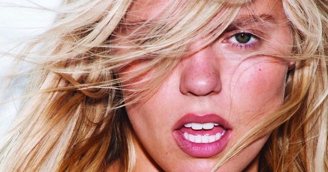 Close-up of a blonde woman with windswept hair, green eyes, pink lips, and sun-kissed skin gazing intensely at the camera.