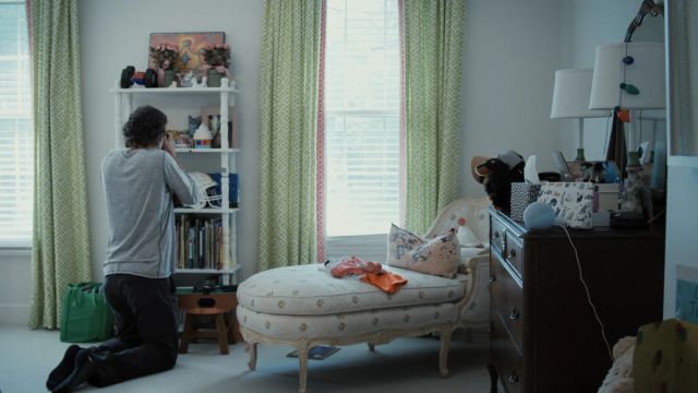 Person kneeling on floor photographing items on a white bookshelf in a bedroom with green curtains, chaise lounge, and dresser.