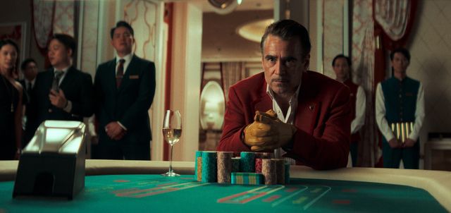 Man in red blazer and leather gloves sits at a casino baccarat table with chips and champagne, staff watching behind him.