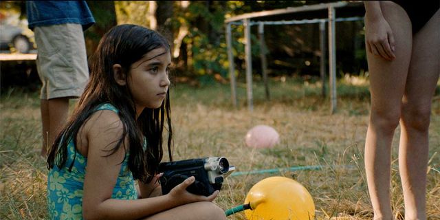 Young girl in blue floral swimsuit sits on grass holding a handheld video camera, with balloons and other people nearby.