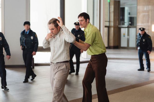 Two men in 1970s-style clothing struggle in a building lobby as police officers in blue uniforms approach from behind.