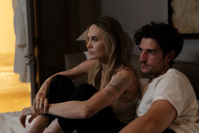 Two people sit close together on a bed in a dimly lit room; a tattooed woman in black and a man in a white t-shirt.