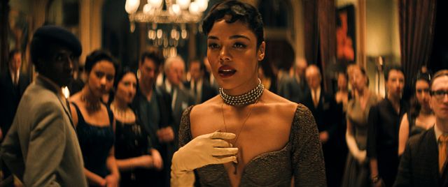 Woman in elegant dark gown and pearl choker with white gloves at a formal gathering, chandelier and crowd behind her.