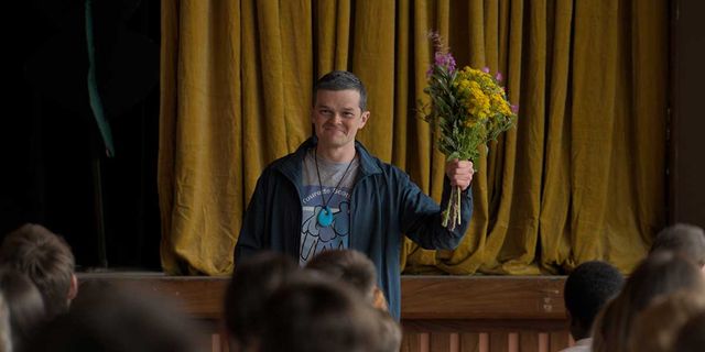 Man smiling on stage holding a bouquet of yellow and purple wildflowers, wearing a blue jacket, with gold curtains behind him.