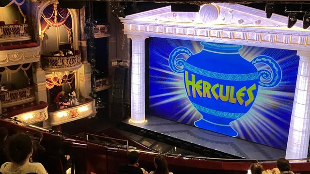 Stage of Hercules musical in an ornate theater, showing a large blue Greek vase with "HERCULES" text, viewed from upper balcony.