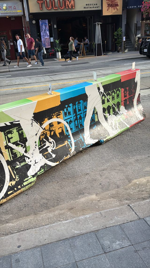 Colorful painted concrete barrier on a city street near Tulum Mexican Restaurant, with pedestrians walking on the sidewalk.