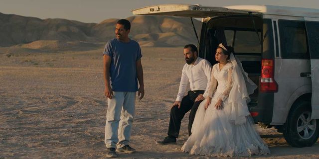 Three people in a desert: a man in blue shirt standing, a couple in wedding attire sitting in the back of a white van.