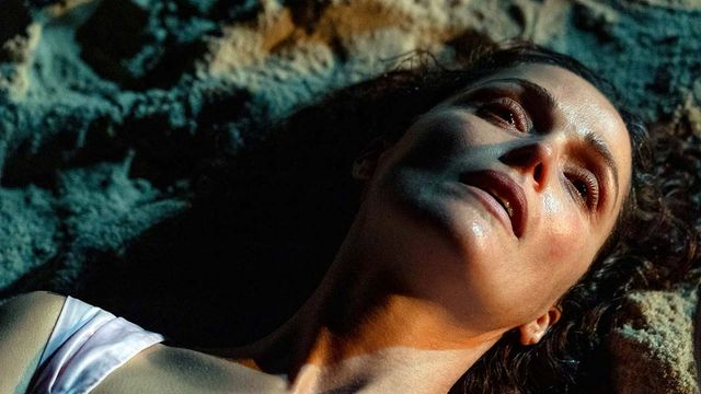 Woman with dark curly hair lying on sand, eyes partially closed, face half in shadow, wearing a white top, dramatic lighting.