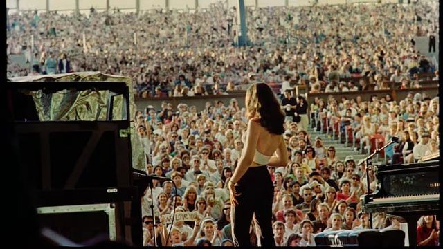 Performer with long dark hair in white top and black pants stands on stage facing a massive outdoor stadium crowd.