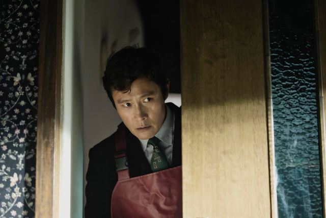 Man in suit with green tie and red apron peers cautiously through a partially open wooden door with a tense expression.
