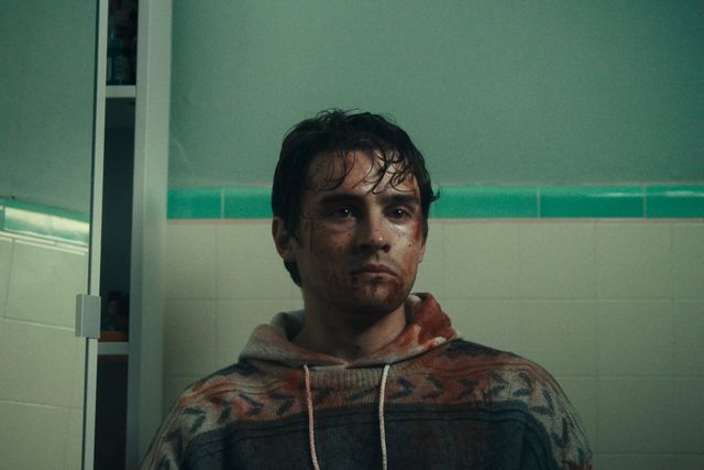 Young man with disheveled wet hair and blood-smeared face wearing a patterned hoodie, sitting in a tiled bathroom.