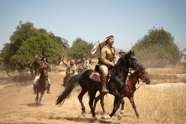 Horseback riders in Middle Eastern attire galloping through dry grassland, carrying rifles, appearing to be a film or reenactment scene.